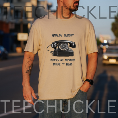A man wearing a sand graphic t-shirt with a black rotary telephone design and the text 'ANALOGUE MEMORY MEMORIZING NUMBERS INSIDE MY HEAD'. The photo is taken outdoors on a street at sunset, 