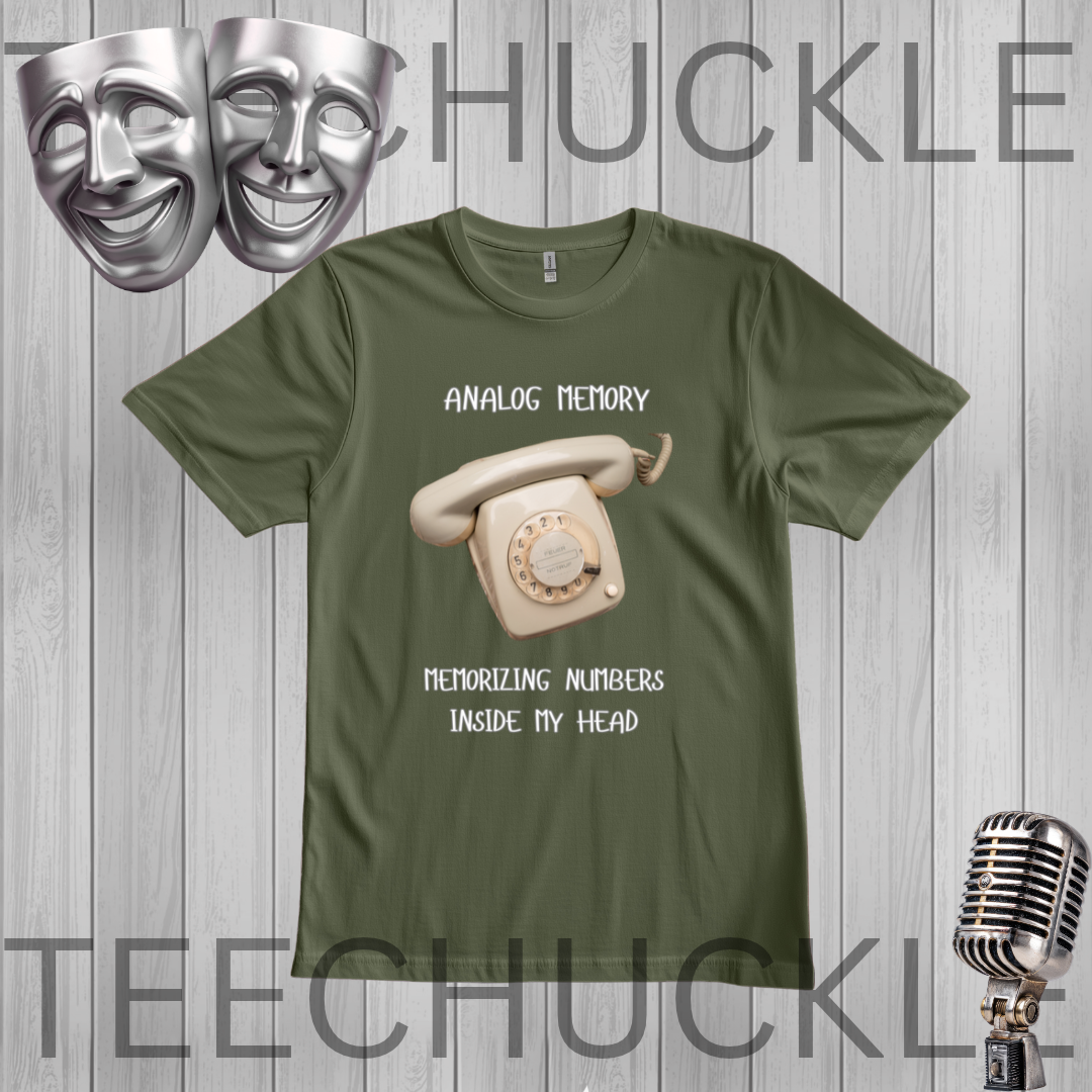 A green graphic t-shirt featuring a vintage cream rotary phone and the text 'ANALOGIC MEMORY' and 'MEMORIZING NUMBERS INSIDE MY HEAD'. The t-shirt is displayed on a gray wood-plank background with a silver microphone, theater masks,