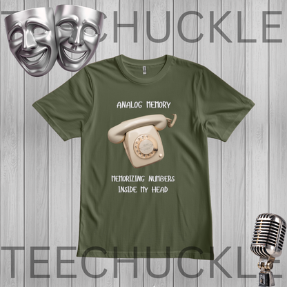 A green graphic t-shirt featuring a vintage cream rotary phone and the text 'ANALOGIC MEMORY' and 'MEMORIZING NUMBERS INSIDE MY HEAD'. The t-shirt is displayed on a gray wood-plank background with a silver microphone, theater masks,