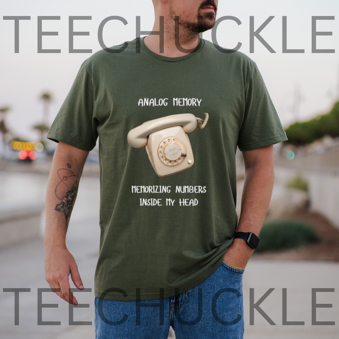 A man wearing a green graphic t-shirt with a retro rotary phone design and the text 'ANALOGIC MEMORY' and 'MEMORIZING NUMBERS INSIDE MY HEAD'.  and the background is a blurred outdoor street scene.