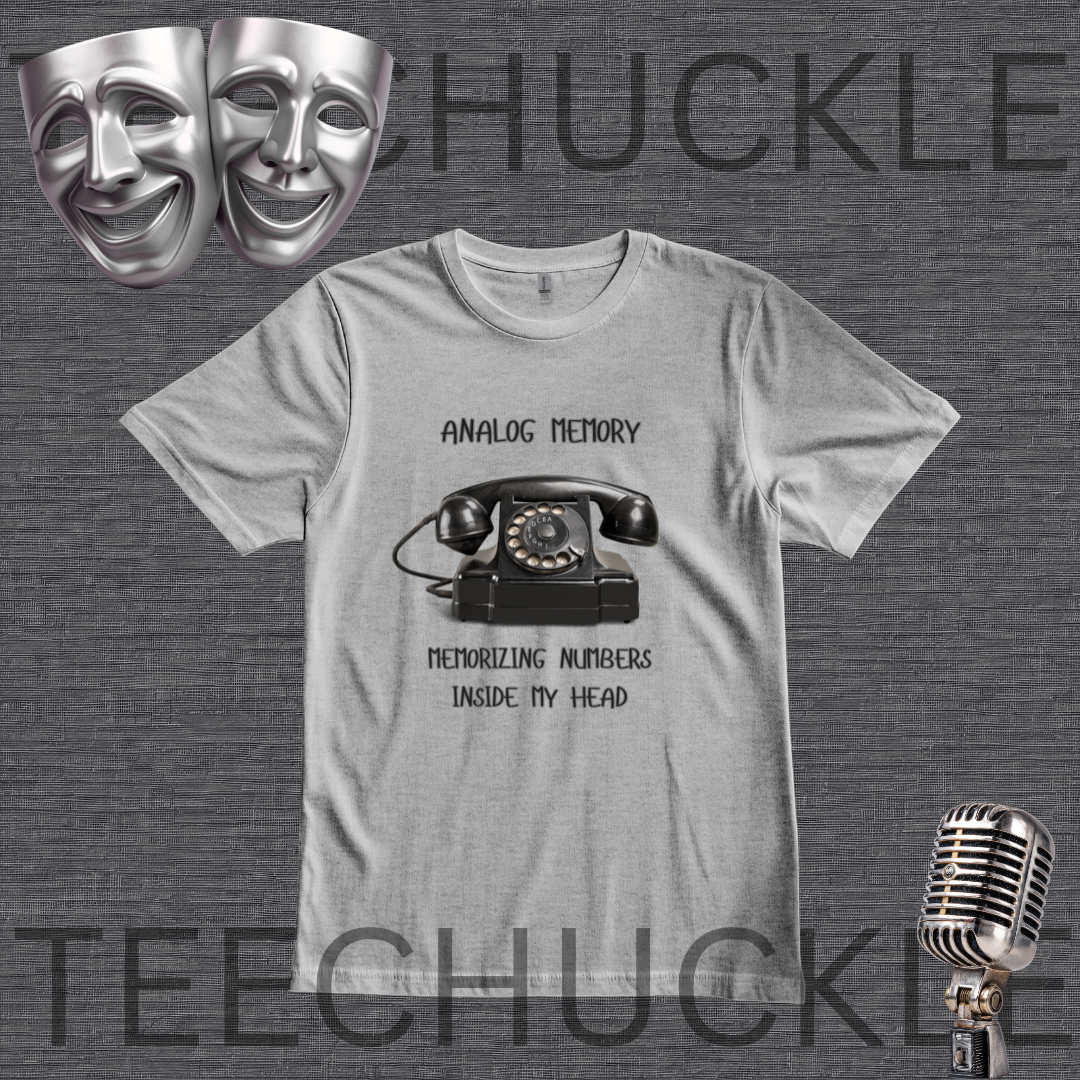 A gray graphic t-shirt featuring a black retro rotary phone and the text 'ANALOG MEMORY' and 'MEMORIZING NUMBERS INSIDE MY HEAD'. The t-shirt is displayed on a dark gray textured background with  decorative silver comedy and tragedy masks and a vintage microphone.