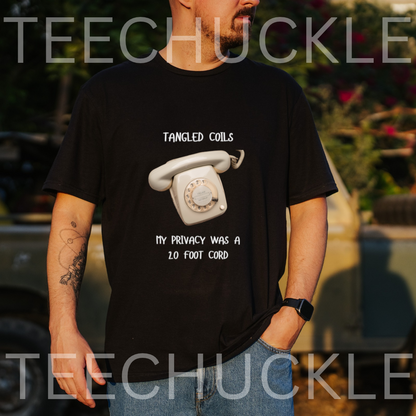 A man wearing a black graphic t-shirt with a white retro rotary phone and the text 'TANGLED COILS MY PRIVACY WAS A 10 FOOT CORD'. The background is an outdoor blurred setting with a vehicle
