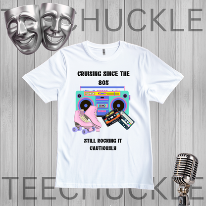 A white graphic t-shirt displayed on a gray wood plank background, featuring a retro 80s design with a boombox, pink roller skate, cassette tapes, and the text 'CRUISING SINCE THE 80S STILL ROCKING IT CAUTIOUSLY'. The image includes decorative silver theater masks and a vintage microphone