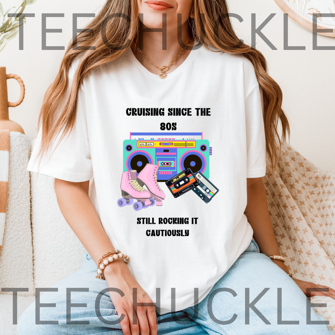 A woman wearing a white graphic t-shirt with a retro 80s theme. The shirt features a colorful boombox, cassette tapes, and a pink roller skate with the text 'CRUISING SINCE THE 80S STILL ROCKING IT CAUTIOUSLY'.