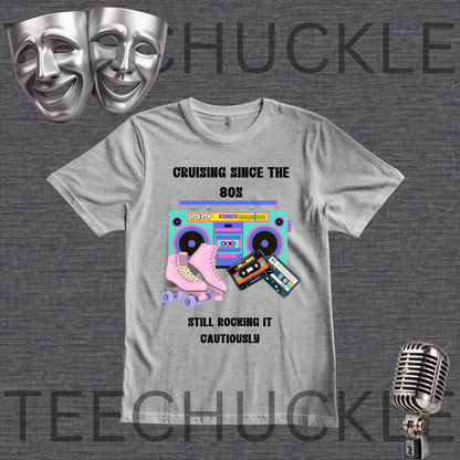 A light gray graphic t-shirt featuring a retro 80s theme with a boombox, roller skate, and cassette tapes, accompanied by the text 'CRUISING SINCE THE 80S STILL ROCKING IT CAUTIONLY'. The t-shirt is displayed on a dark gray textured background with silver theater masks, a vintage microphone