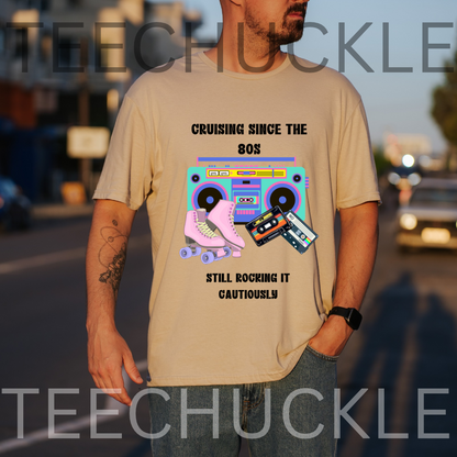 A man wearing a sand graphic t-shirt with a retro 80s boombox and roller skate design, standing outdoors on a blurred street background. The t-shirt features the text 'CRUISING SINCE THE 80S' and 'STILL ROCKING IT CAUTIOUSLY'.