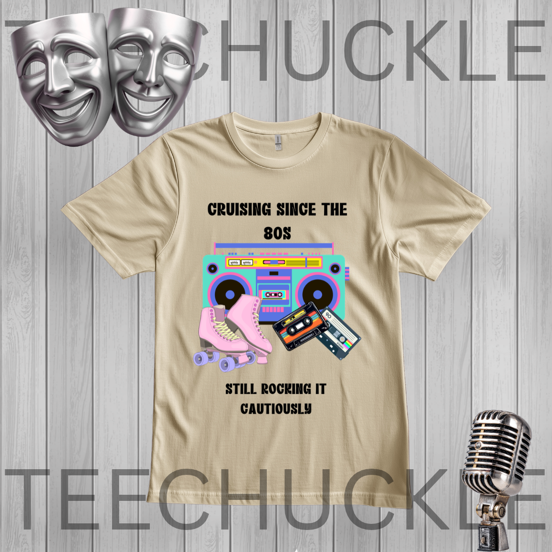 A sand graphic t-shirt featuring retro 80s-themed illustrations including a pink roller skate, a pink boombox, cassette tapes, and the text 'CRUISING SINCE THE 80s STILL ROCKING IT CAUTIONARILY'. The t-shirt is displayed on a gray wood-plank background with silver theater masks, a vintage microphone
