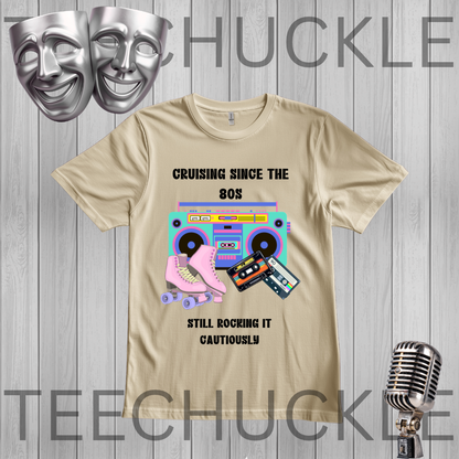 A sand graphic t-shirt featuring retro 80s-themed illustrations including a pink roller skate, a pink boombox, cassette tapes, and the text 'CRUISING SINCE THE 80s STILL ROCKING IT CAUTIONARILY'. The t-shirt is displayed on a gray wood-plank background with silver theater masks, a vintage microphone