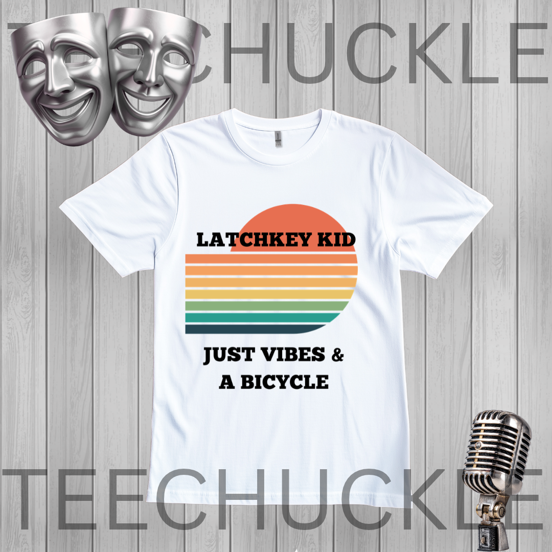 A white graphic t-shirt displayed on a gray wood-plank background, featuring a retro sunset design with the text 'LATCHKEY KID JUST VIBES & A BICYCLE'. The image includes decorative props like silver theater masks and a vintage microphone