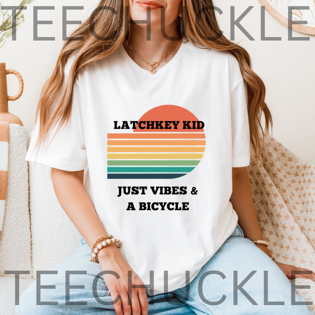 A white graphic t-shirt, featuring a retro sunset design with the text 'LATCHKEY KID JUST VIBES & A BICYCLE'. 