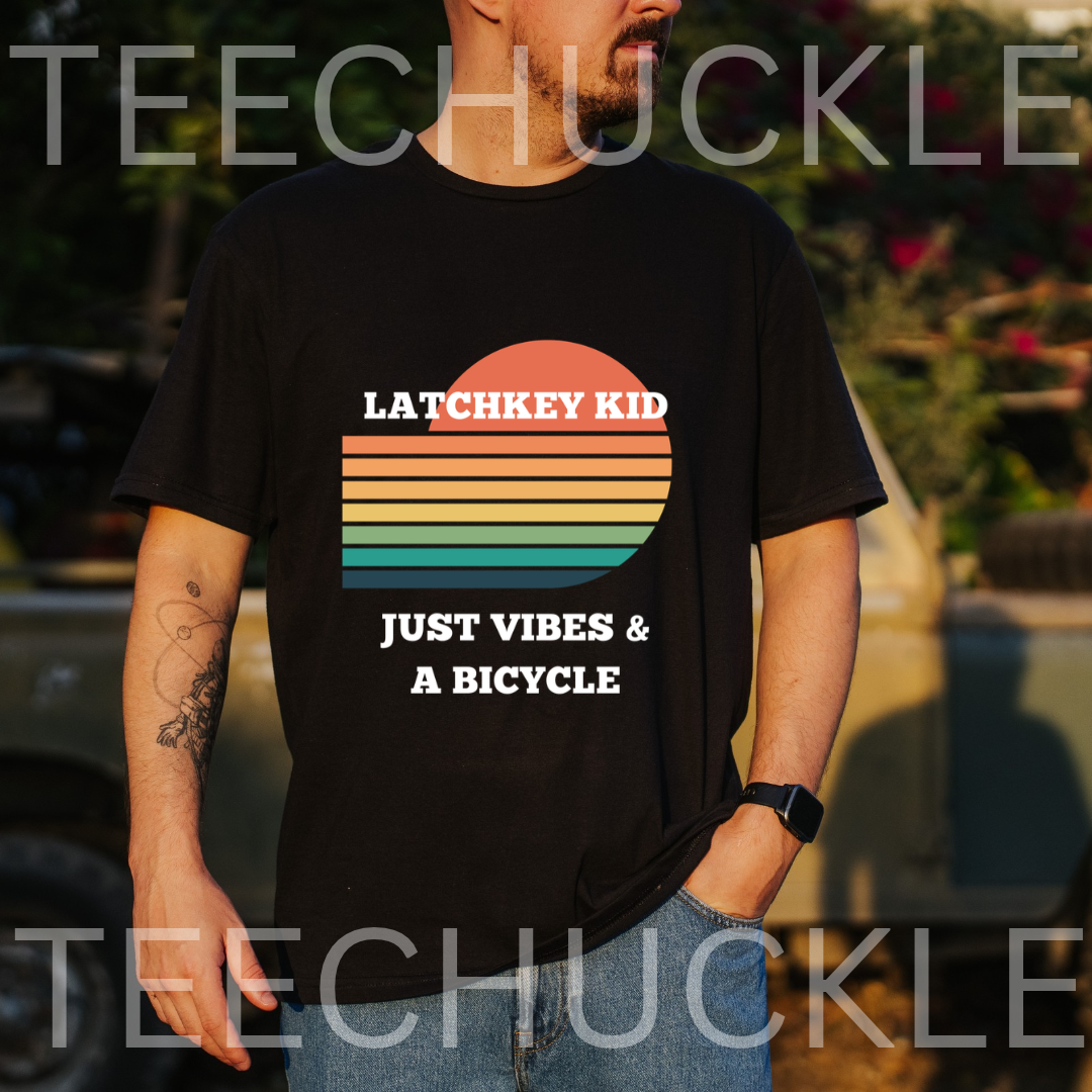 A man wearing a black graphic t-shirt with a retro sunset design and the text 'LATCHKEY KID JUST VIBES & A BICYCLE'. The background is a blurred outdoor setting with warm sunset lighting.