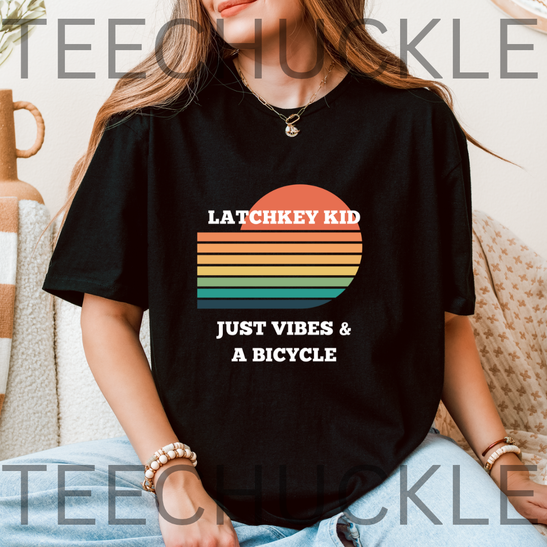 A woman wearing a black graphic t-shirt with a retro sunset design and the text 'LATCHKEY KID JUST VIBES & A BICYCLE'. 