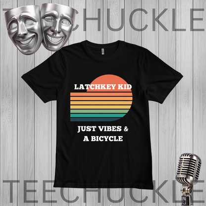 A black short-sleeve graphic T-shirt displayed on a gray wood-plank background, with a retro sunset stripe design in orange, yellow, beige, green, and blue, plus white text reading 'LATCHKEY KID JUST VIBES & A BICYCLE.' Silver theater masks and a vintage microphone appear as decorative props