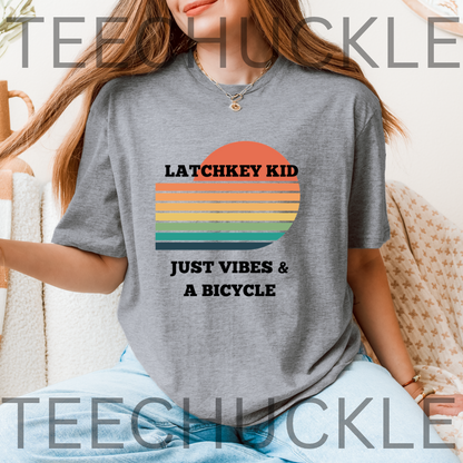 A woman wearing a gray graphic t-shirt with a retro sunset design and the text 'LATCHKEY KID JUST VIBES & A BICYCLE'.