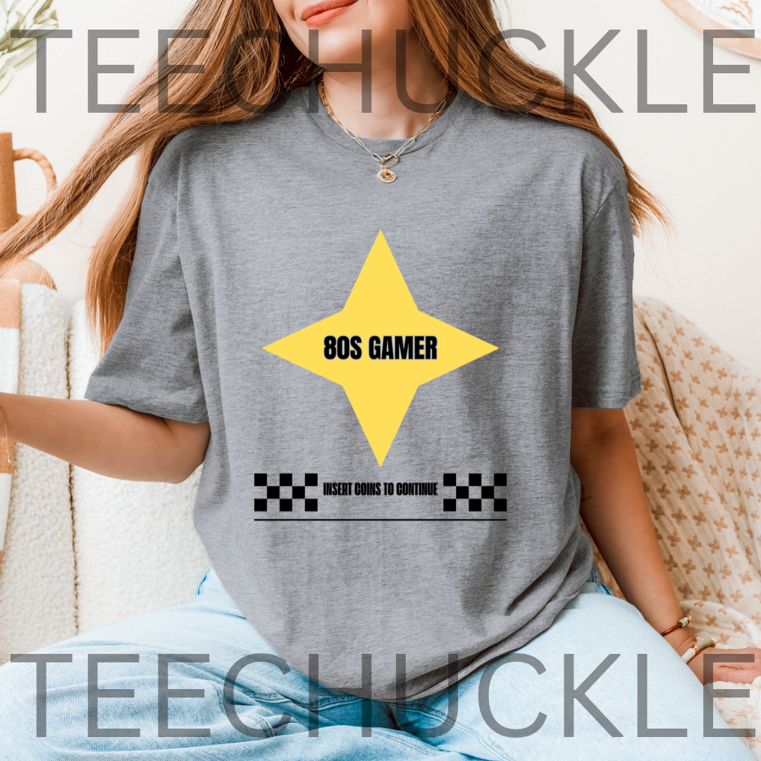 A woman wearing a heather gray t-shirt with a yellow star graphic and the text '80S GAMER' and 'INSERT COIN TO CONTINUE' on the front. 