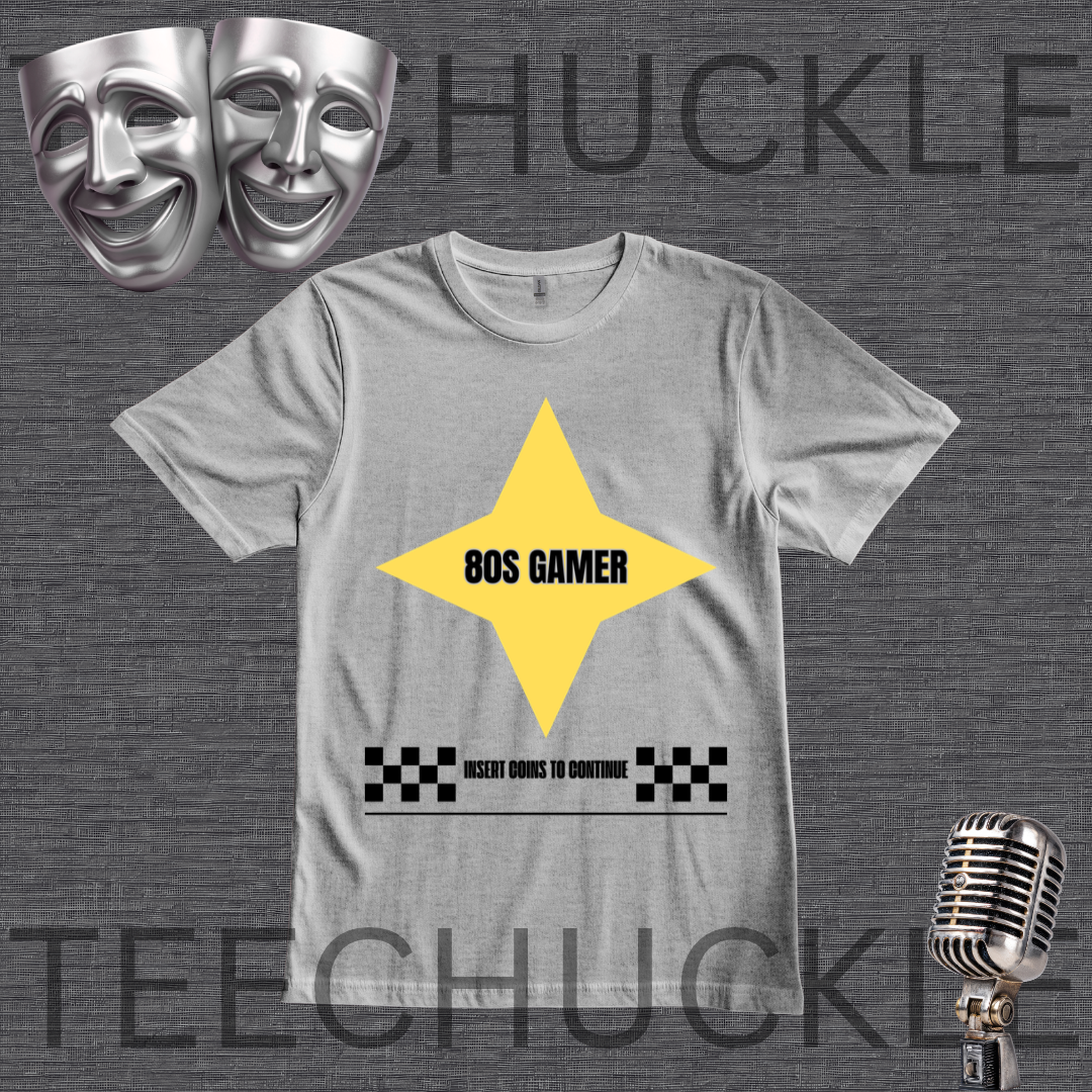 A gray crewneck t-shirt with a graphic design featuring a yellow diamond with '80S GAMER' in black, a black-and-white checkered bar, and placeholder text. The image is set on a dark gray textured background with decorative props including silver comedy and tragedy masks and a vintage microphone.
