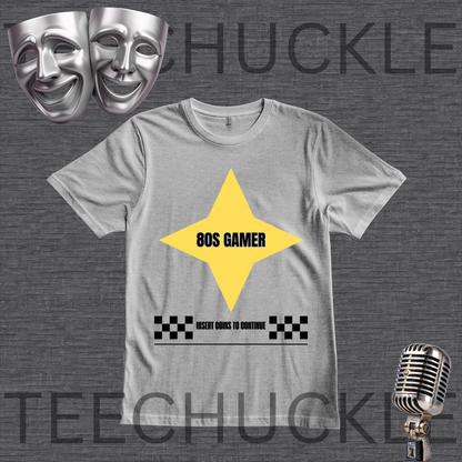 A gray crewneck t-shirt with a graphic design featuring a yellow diamond with '80S GAMER' in black, a black-and-white checkered bar, and placeholder text. The image is set on a dark gray textured background with decorative props including silver comedy and tragedy masks and a vintage microphone.