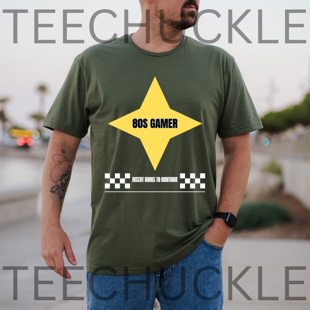 A man wearing a green t-shirt with a yellow star graphic that reads '80S GAMER' and a white checkered bar graphic below it. The background is a blurred outdoor street scene
