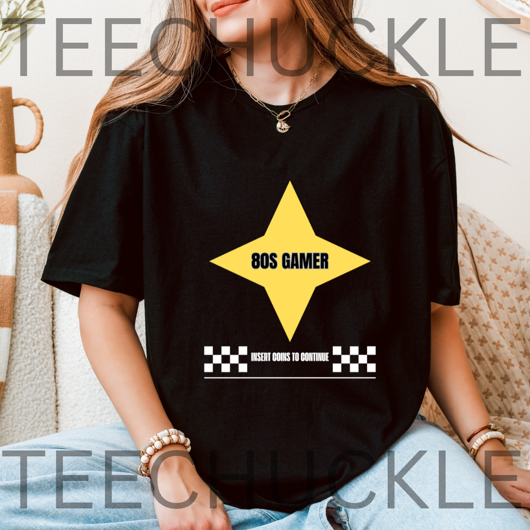 A black short-sleeve graphic t-shirt  The shirt features a large yellow star graphic with the text '80S GAMER' in black, and a white text bar below reading 'INSERT COINS TO CONTINUE' flanked by black-and-white checkered squares.