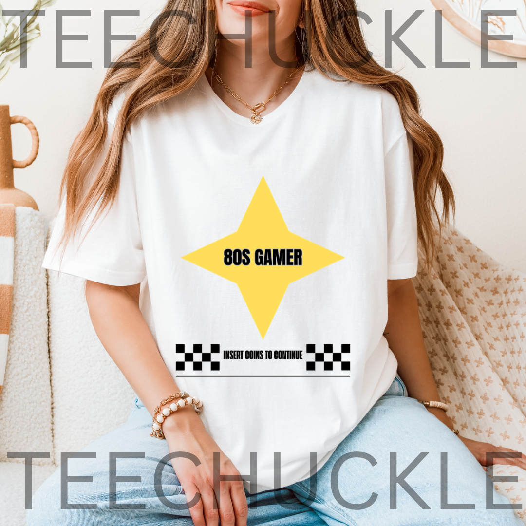 A woman wearing a white graphic t-shirt with a yellow star and '80S GAMER' text, sitting in a cozy indoor setting. 