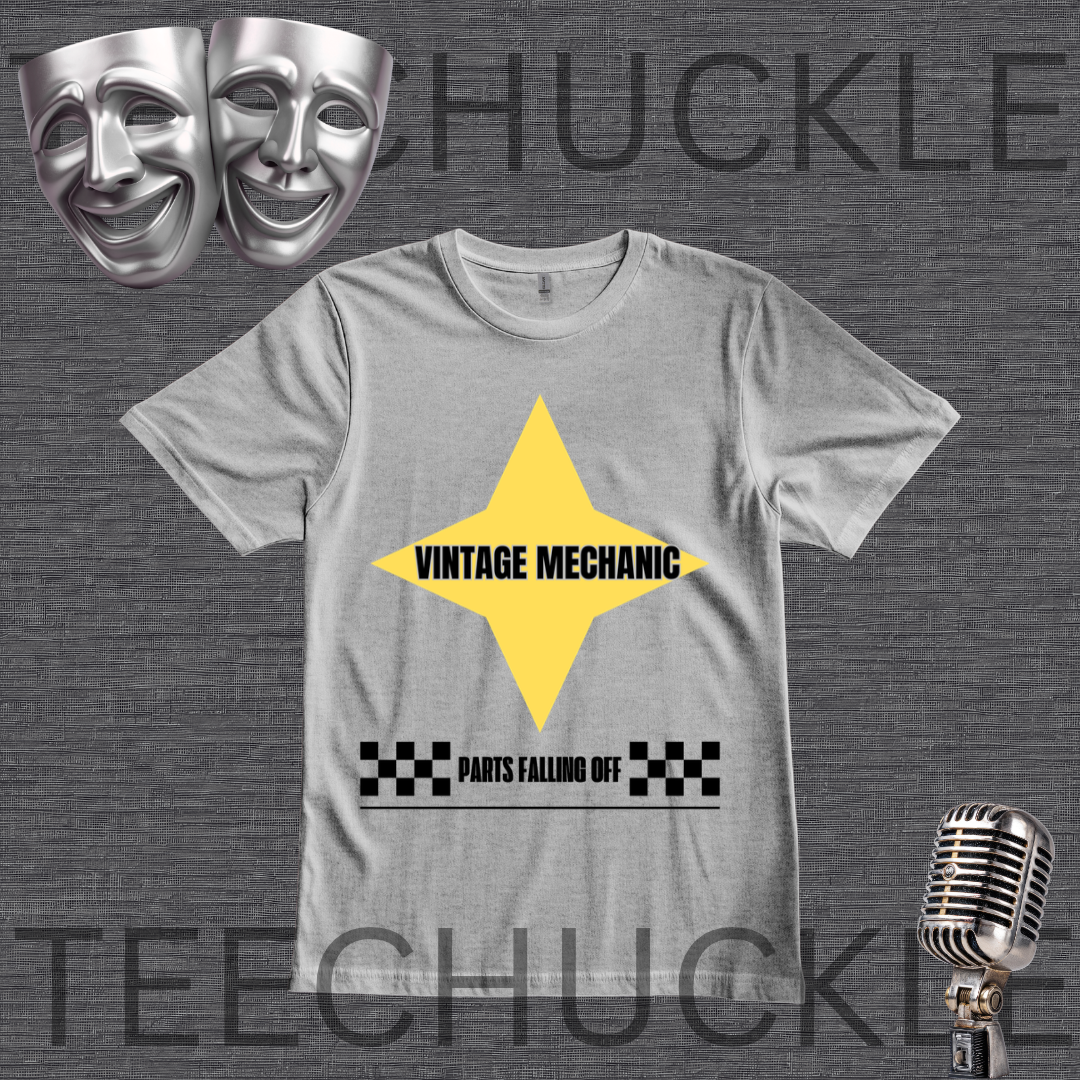A gray crewneck t-shirt featuring a yellow diamond graphic with the text 'VINTAGE MECHANIC' and a black-and-white checkered bar below reading 'PARTS FALLING OFF'. The shirt is displayed on a dark gray textured background and decorative silver comedy and microphone masks.