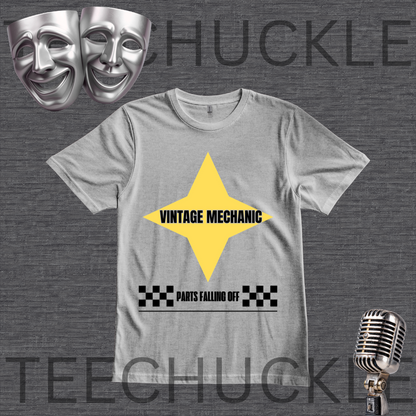 A gray crewneck t-shirt featuring a yellow diamond graphic with the text 'VINTAGE MECHANIC' and a black-and-white checkered bar below reading 'PARTS FALLING OFF'. The shirt is displayed on a dark gray textured background and decorative silver comedy and microphone masks.