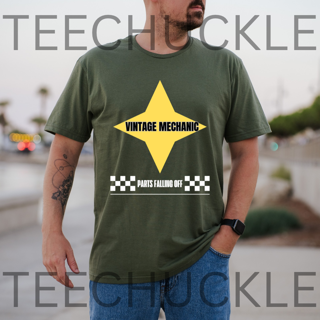 A man wearing a green t-shirt with a yellow star graphic that reads ' 'VINTAGE MECHANIC' in black, plus a black-and-white checkered bar and the phrase 'PARTS FALLING OFF' below. The background is a blurred outdoor street scene