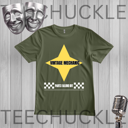 A green graphic t-shirt featuring a yellow star with 'VINTAGE MECHANIC' in black text and a white checkered bar below reading 'PARTS FALLING OFF'. The t-shirt is displayed on a gray wood-plank background  flanked by silver comedy and tragedy masks on the top left and a vintage microphone on the bottom right.