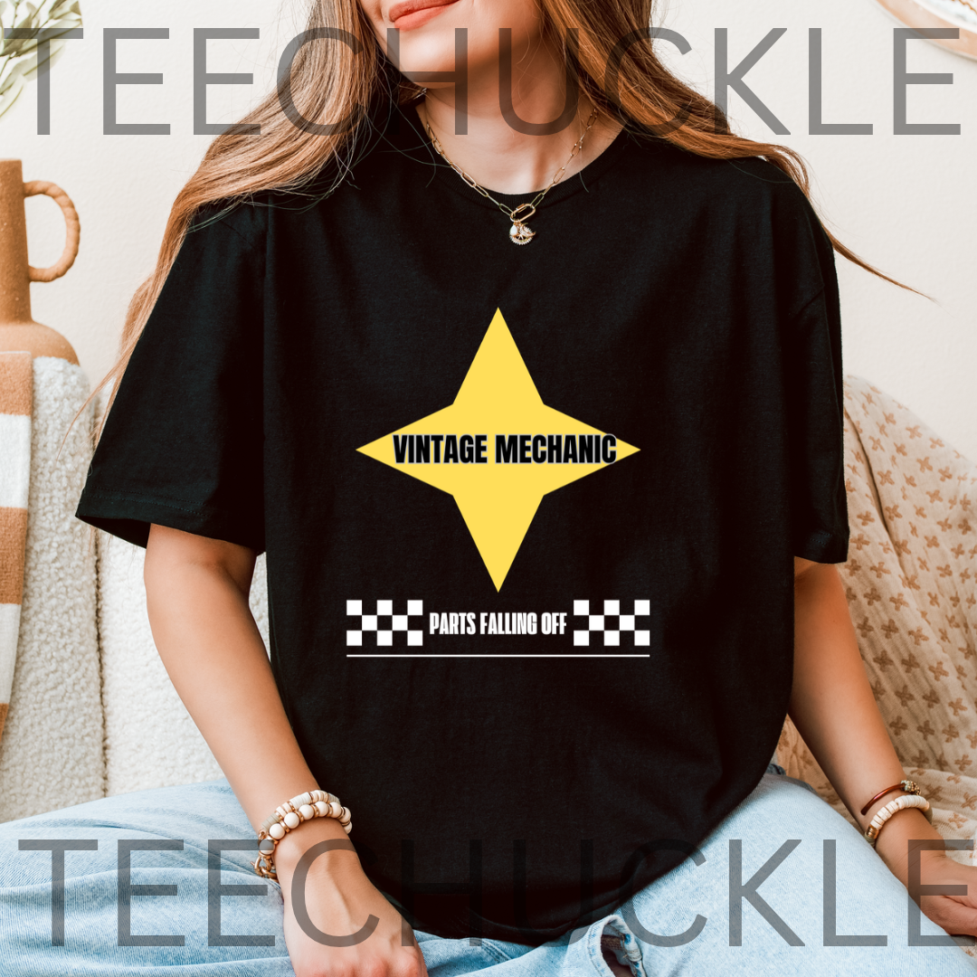 A woman wearing a black graphic T-shirt with a yellow star and the words 'VINTAGE MECHANIC' and 'PARTS FALLING OFF' on the front, seated indoors with a neutral background
