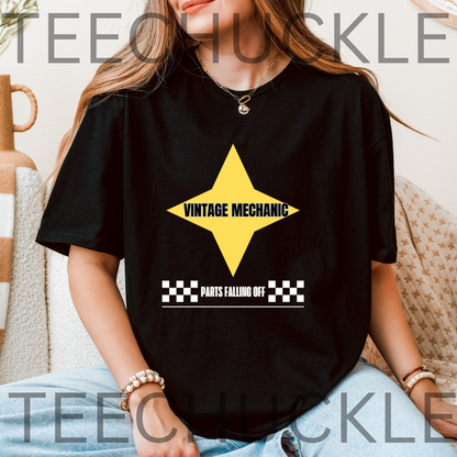 A woman wearing a black graphic T-shirt with a yellow star and the words 'VINTAGE MECHANIC' and 'PARTS FALLING OFF' on the front, seated indoors with a neutral background