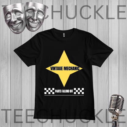 A black short-sleeve graphic t-shirt featuring a yellow star with 'VINTAGE MECHANIC' in black text, and a white checkered bar with 'PARTS FALLING OFF' underneath. The shirt is displayed on a gray wood-plank background with a vintage microphone, silver comedy and tragedy masks
