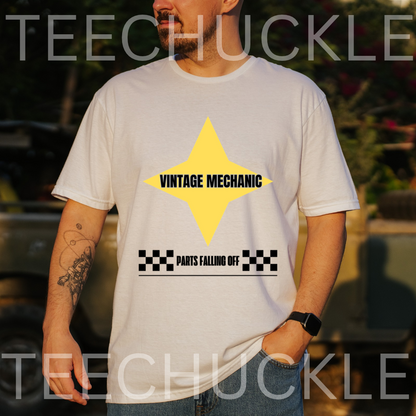 A man wearing a white graphic t-shirt with a yellow star and black text reading 'VINTAGE MECHANIC' and 'PARTS FALLING OFF'. The background is a blurred outdoor setting with a vintage car, 