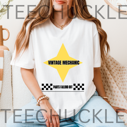 A woman wearing a white graphic T-shirt with a yellow star and black text reading 'VINTAGE MECHANIC' and 'PARTS FALLING OFF', seated indoors on a patterned couch. 