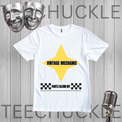 A white short-sleeve graphic T-shirt laid flat on a gray wood-plank background. The shirt has a large yellow star shape with the words 'VINTAGE MECHANIC' in black, plus a black-and-white checkered bar and the phrase 'PARTS FALLING OFF' below.
