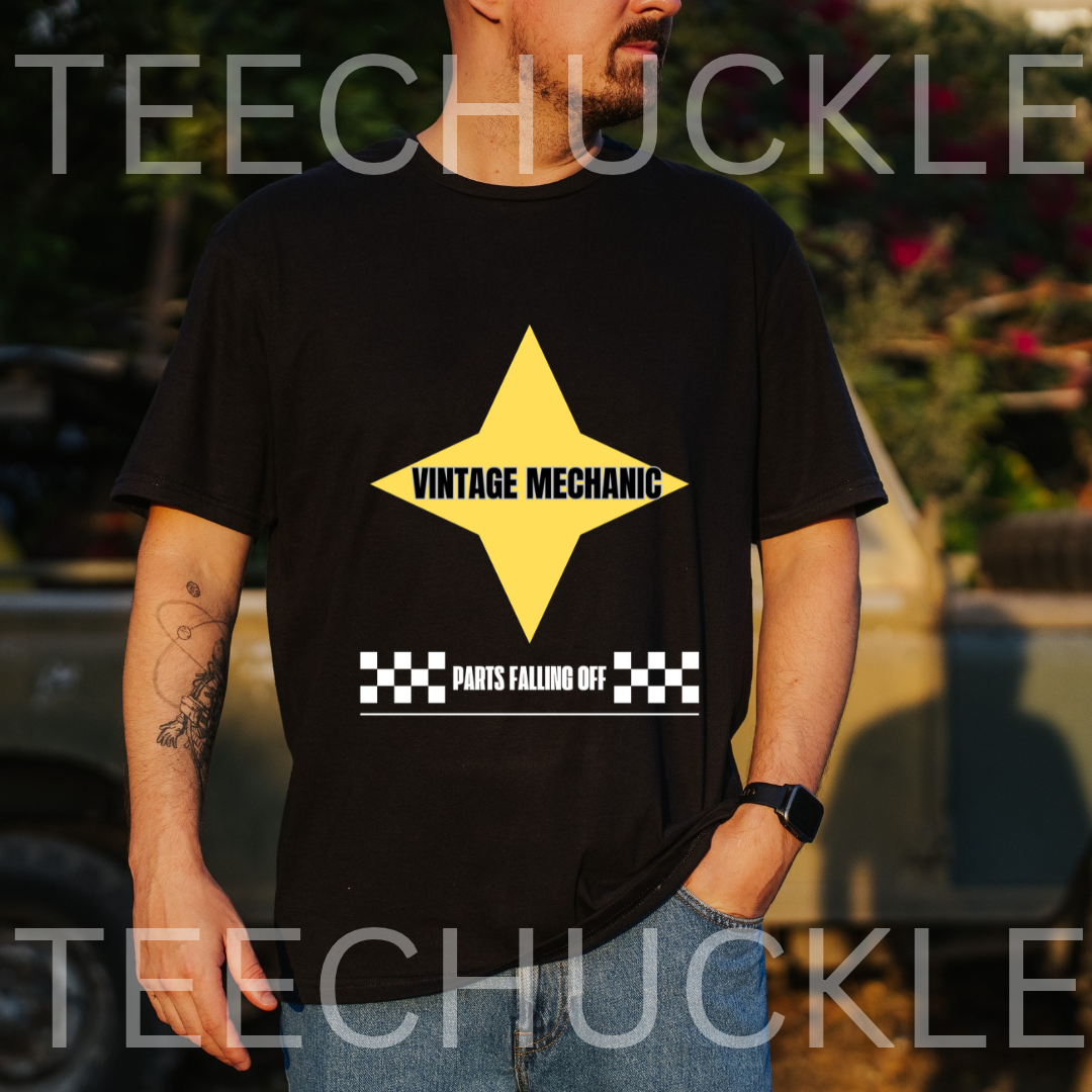 A man wearing a black graphic t-shirt with a yellow diamond logo that reads 'VINTAGE MECHANIC' and a white banner below reading 'PARTS FALLING OFF' with a black-and-white checkered pattern. The background is an outdoor setting with a blurred vehicle and foliage, 