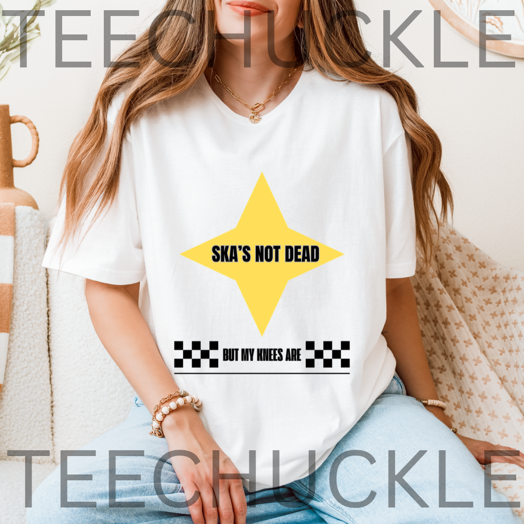 A woman wearing a white graphic t-shirt with a yellow star and the text 'SKA'S NOT DEAD BUT MY KNEES ARE' in black, set against a cozy indoor background 
