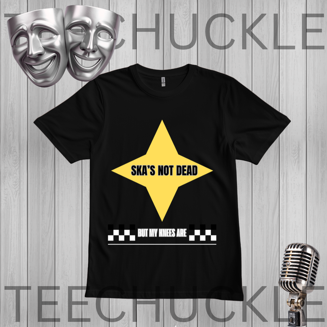 A black short-sleeve graphic t-shirt featuring a yellow star with the text 'SKA'S NOT DEAD' and a white checkerboard bar with the text 'BUT MY WHEELS ARE' underneath. The t-shirt is displayed on a gray wood-plank background with silver theater masks and a vintage microphone as props