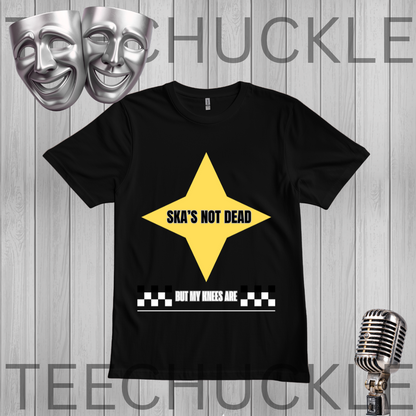 A black short-sleeve graphic t-shirt featuring a yellow star with the text 'SKA'S NOT DEAD' and a white checkerboard bar with the text 'BUT MY WHEELS ARE' underneath. The t-shirt is displayed on a gray wood-plank background with silver theater masks and a vintage microphone as props