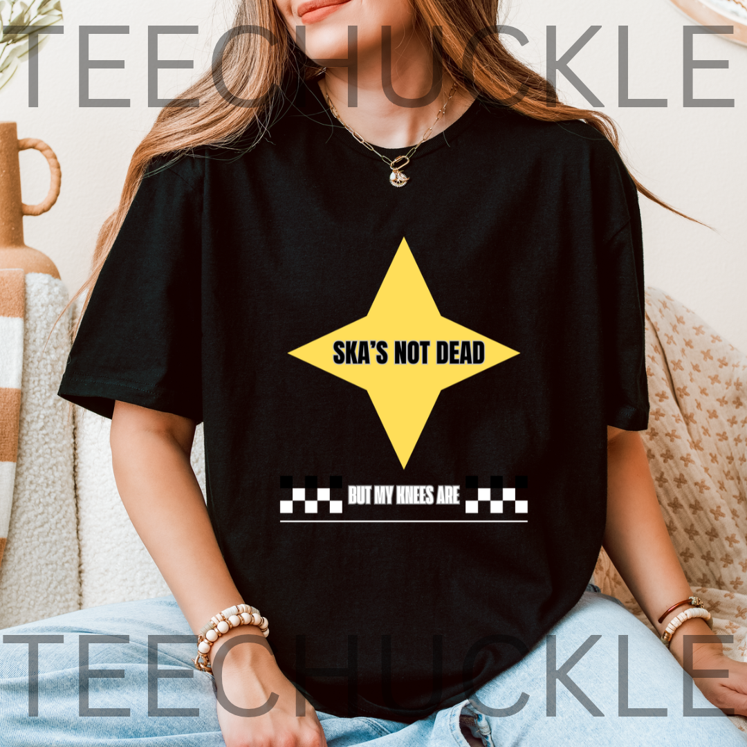 A woman wearing a black graphic t-shirt with a yellow star and the text 'SKA'S NOT DEAD' and 'BUT MY KNEES ARE' on a checkered strip. 