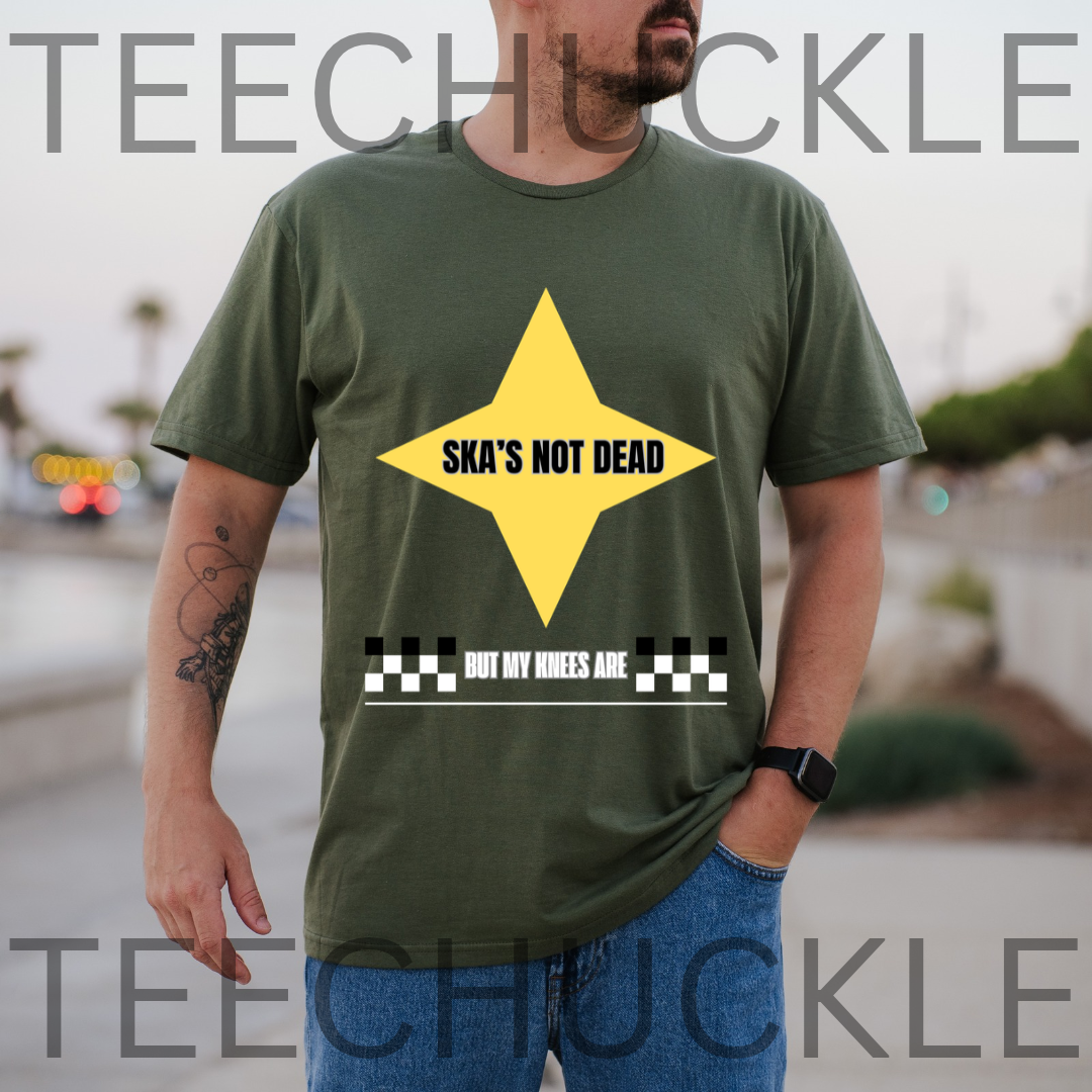 A man wearing a green graphic t-shirt with a yellow diamond and black text reading 'SKA'S NOT DEAD BUT MY WHEELS ARE', and background is a blurred outdoor street scene.