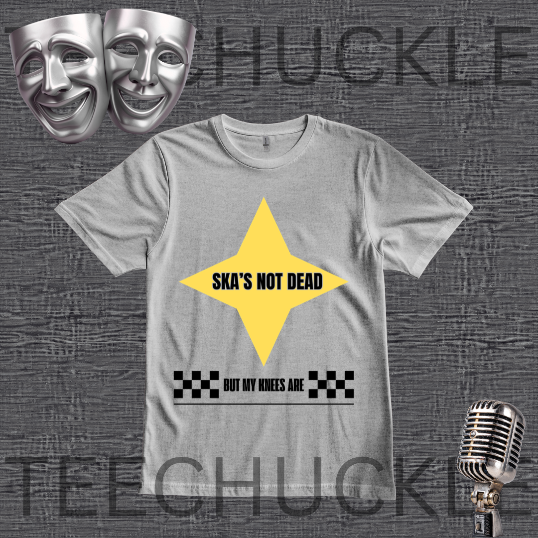 A gray graphic t-shirt featuring a yellow star with the text 'SKA'S NOT DEAD' and a black-and-white checkered pattern with the text 'BUT MY KNEES ARE' underneath. The t-shirt is displayed on a dark gray textured background with a vintage microphone, silver comedy masks