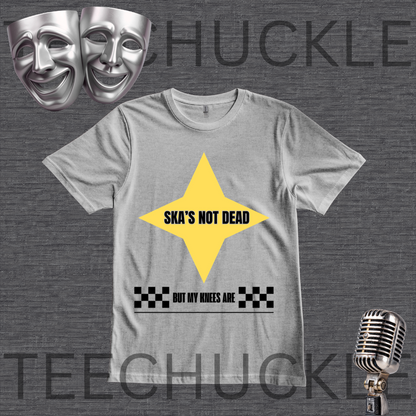 A gray graphic t-shirt featuring a yellow star with the text 'SKA'S NOT DEAD' and a black-and-white checkered pattern with the text 'BUT MY KNEES ARE' underneath. The t-shirt is displayed on a dark gray textured background with a vintage microphone, silver comedy masks