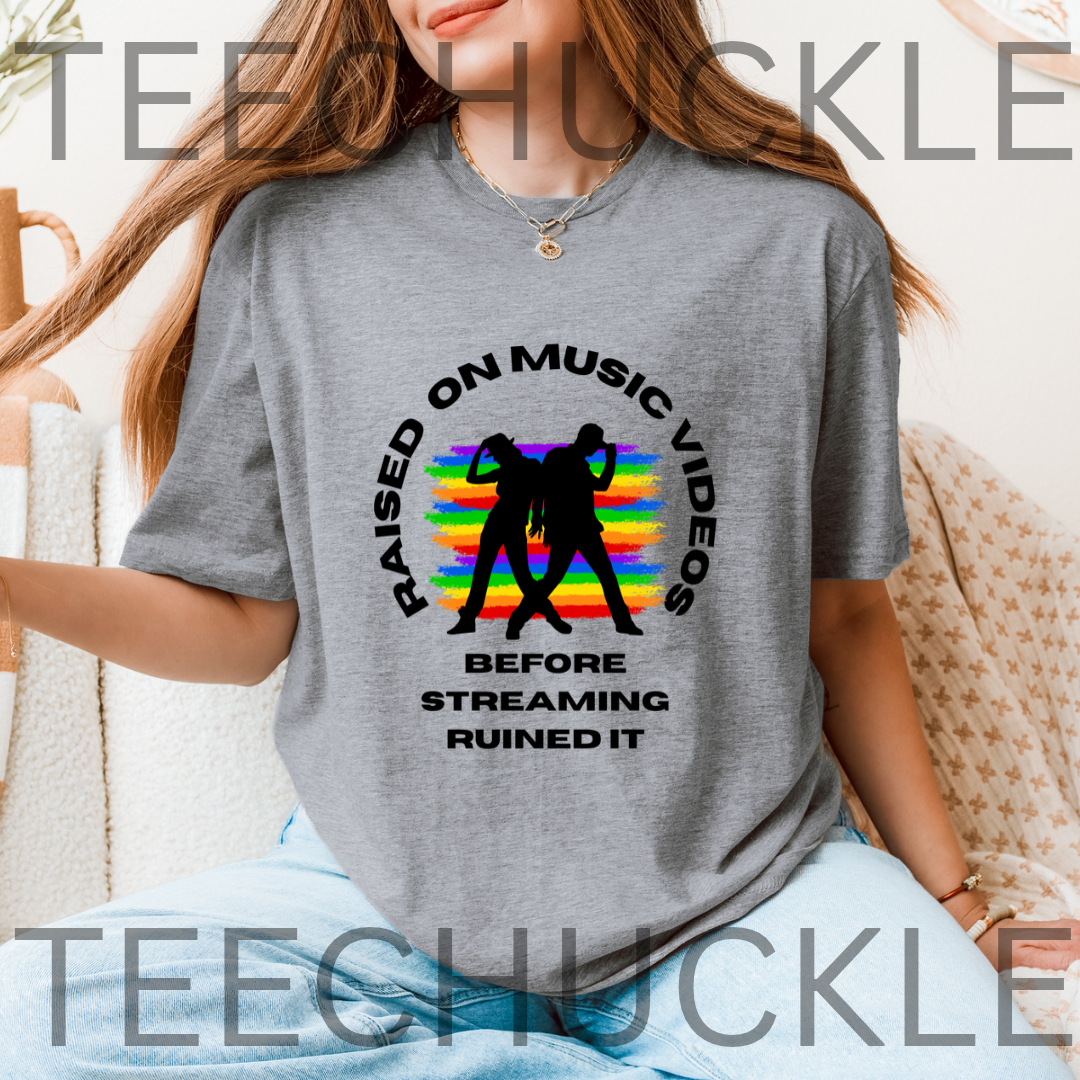 A woman wearing a  gray graphic t-shirt with a black and rainbow design that reads 'RAISED ON MUSIC VIDEOS BEFORE STREAMING RUINED IT'. 