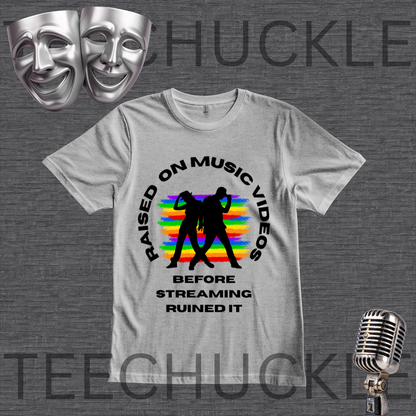 A gray graphic t-shirt displayed on a dark gray textured background. The shirt features a black circular graphic with the text 'RAISED ON MUSIC VIDEOS BEFORE STREAMING RUINED IT' surrounding a black silhouette of a person in front of a colorful rainbow-striped background.