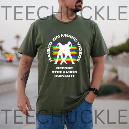 A man wearing a green graphic t-shirt with a retro rainbow design and white text that reads 'RAISED ON MUSIC VIDEOS BEFORE STREAMING RUINED IT'. The background is a blurred outdoor street scene