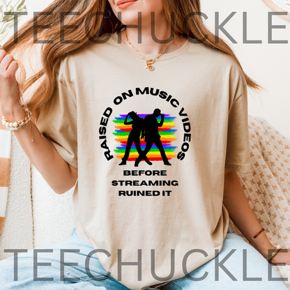 A woman wearing a sand graphic t-shirt with a black circular text design and a rainbow-striped silhouette of two dancing figures. The text reads 'RAISED ON MUSIC VIDEOS BEFORE STREAMING RUINED IT'.