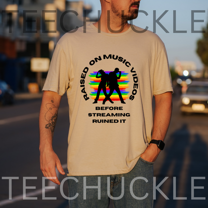 A man wearing a beige graphic t-shirt with a black and rainbow design that reads 'RAISED ON MUSIC VIDEOS BEFORE STREAMING RUINED IT'. The background is a blurred street scene at sunset