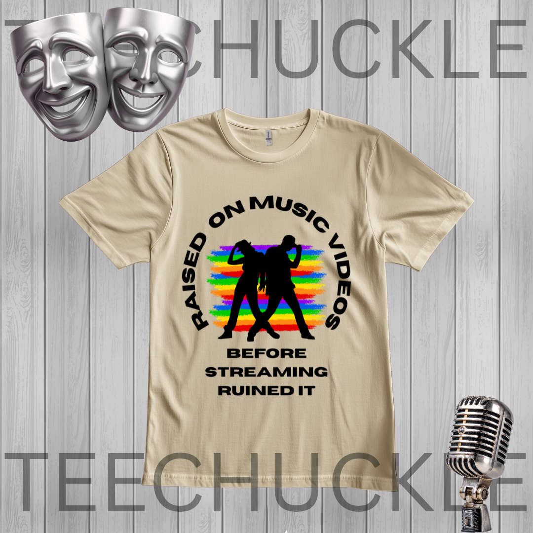 A sand graphic t-shirt displayed on a gray wood-plank background. The shirt features a black circular graphic with the text 'RAISED ON MUSIC VIDEOS BEFORE STREAMING RUINED IT' surrounding a black silhouette of two people dancing in front of a rainbow-striped background.