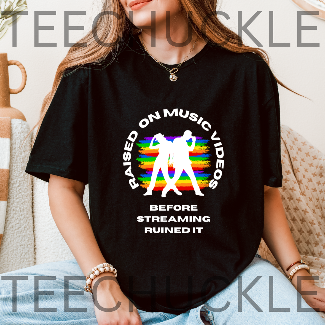 A woman wearing a black graphic t-shirt with a white circular design featuring two figures over a rainbow and the text 'RAISED ON MUSIC VIDEOS BEFORE STREAMING RUINED IT'.