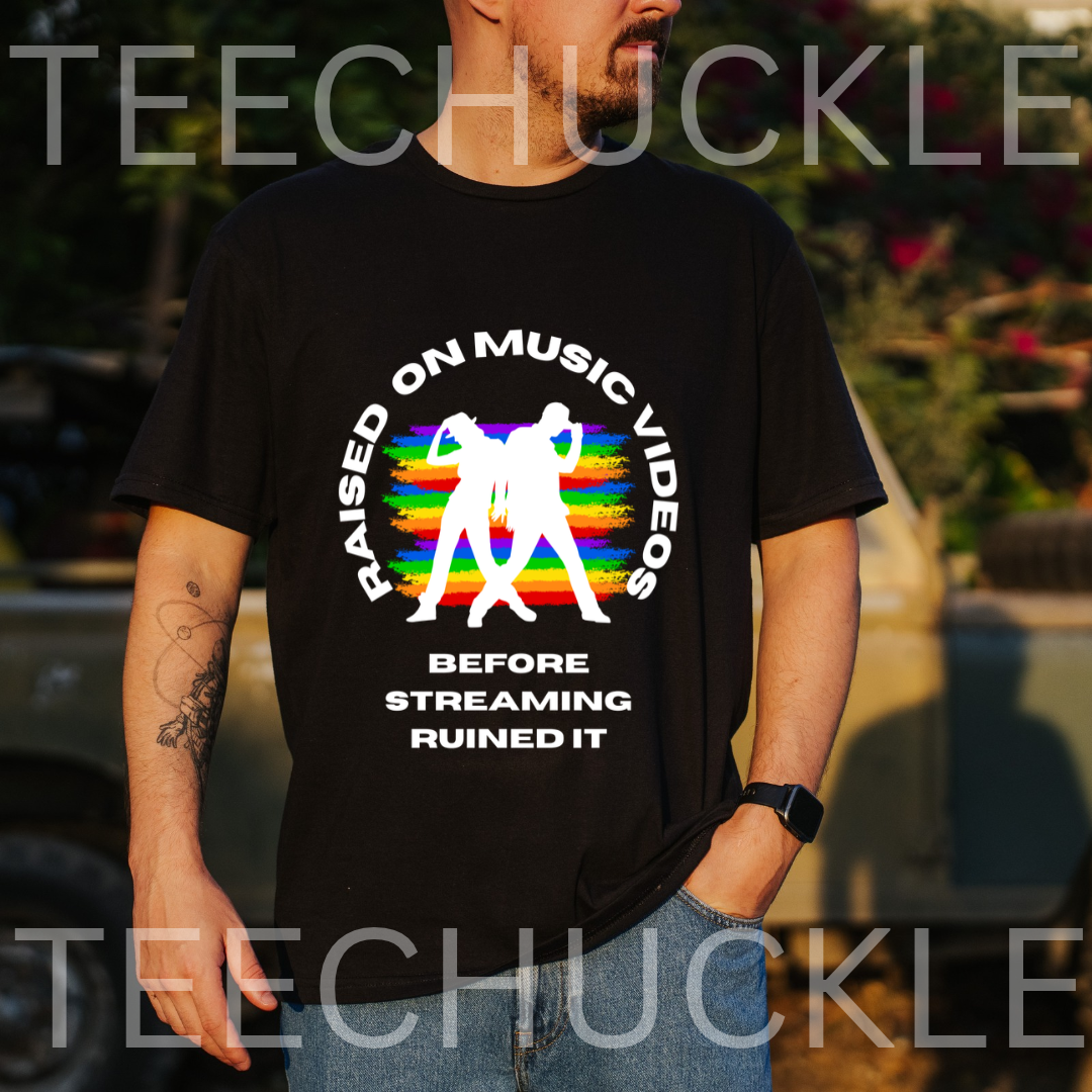 A man wearing a black graphic t-shirt with a colorful rainbow design and white text that reads 'RAISED ON MUSIC VIDEOS BEFORE STREAMING RUINED IT'. The background is an outdoor setting with a blurred vehicle and foliage.